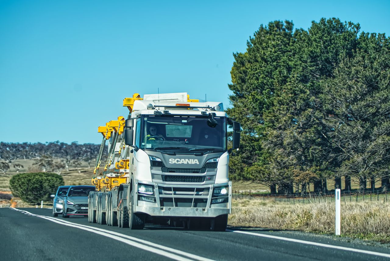 A Scania truck drives down a rural road under a clear blue sky, surrounded by trees.